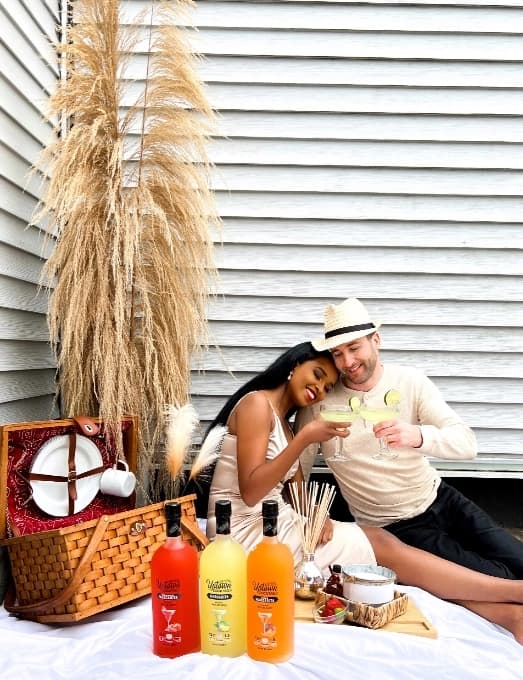Couple clinking margarita glasses at an outdoor picnic with bottled cocktail mixers.