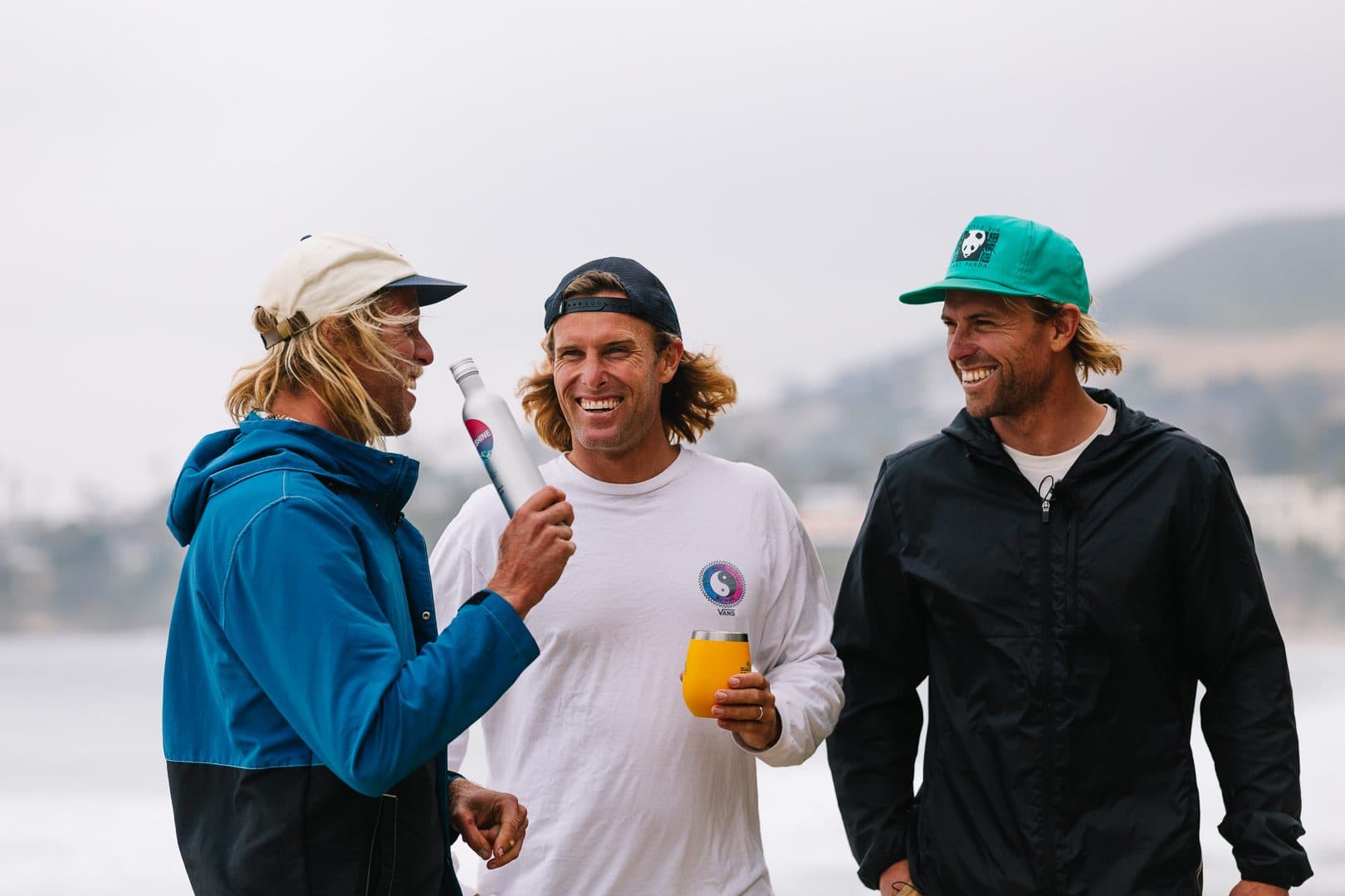 Three men laughing outdoors, one holding a metal beverage bottle and an orange tumbler