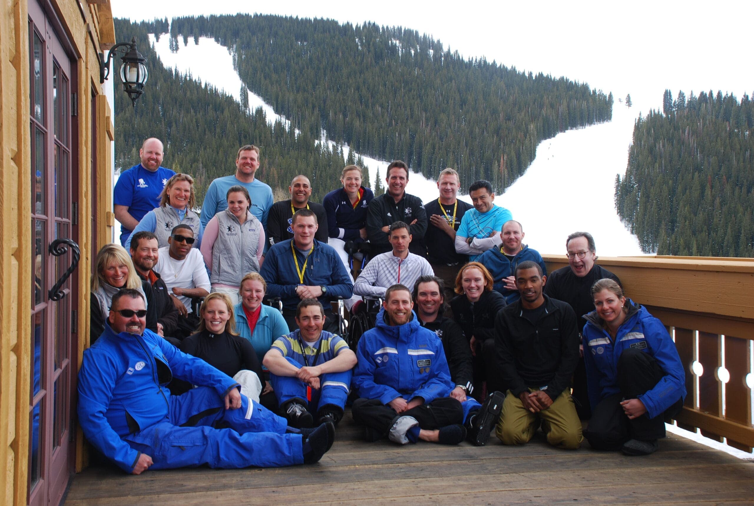 Large group of adaptive skiers and staff pose on a deck overlooking a ski slope